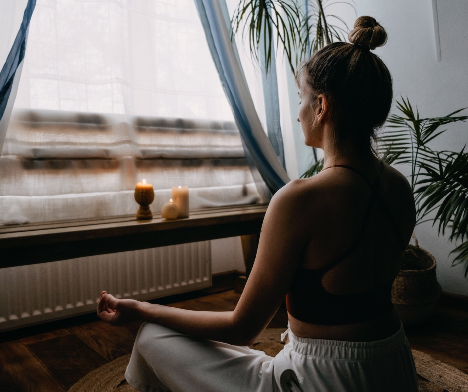 A woman is practicing meditation in a calm, indoor setting, sitting cross-legged on the floor in front of a window. The room is softly lit with natural light, and candles are glowing on a wooden bench in the background. The peaceful ambiance, combined with plants around her, creates a serene and mindful atmosphere, perfect for relaxation and self-care.